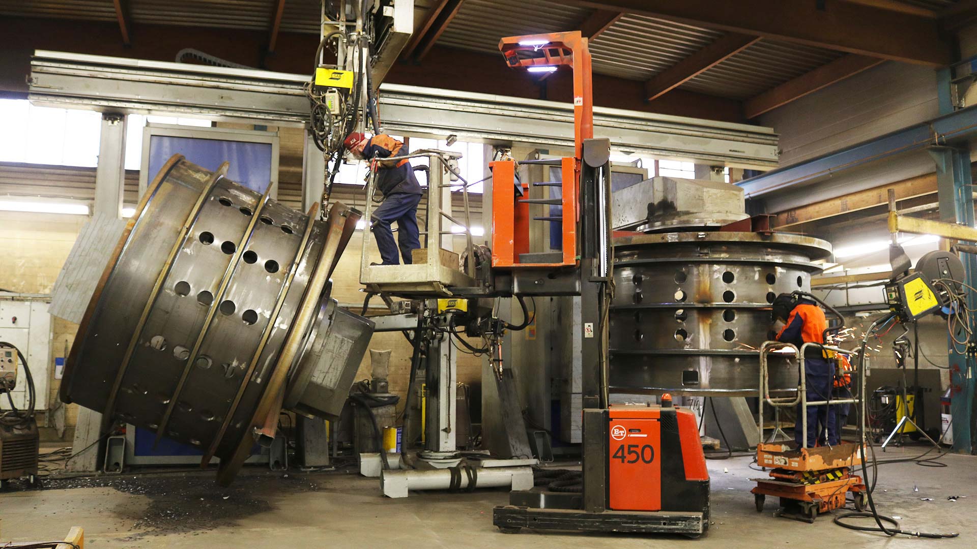 Robotic welding of large cylindrical metal component using lift equipment.